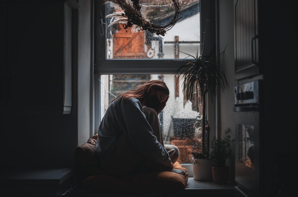 girl sitting in dark window wearing a mask