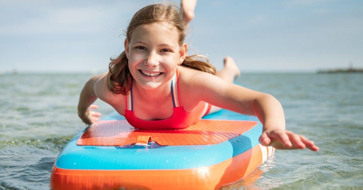 A young girl smiling toward the viewer as she rides on a blue-and-orange paddleboard on a large body of calm, open water.