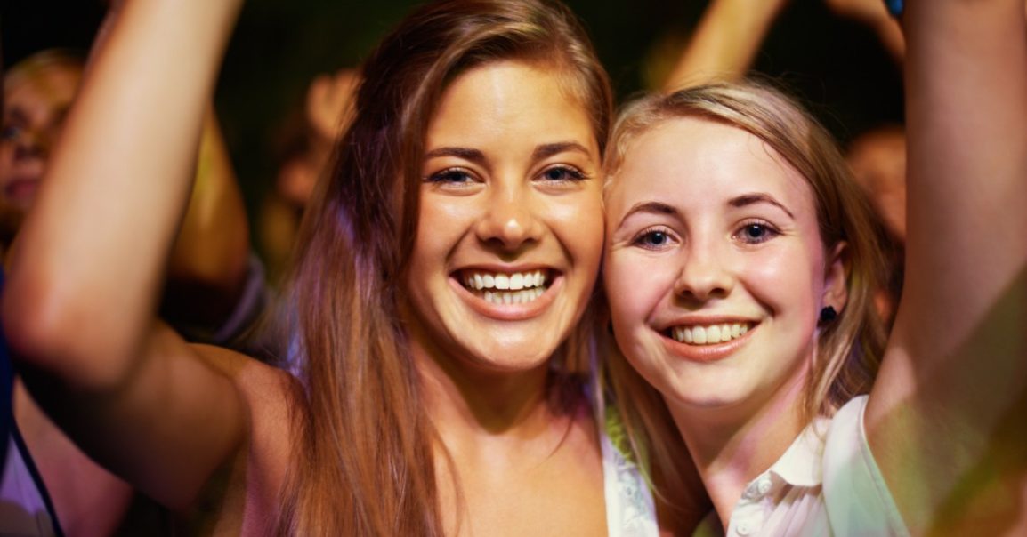Two young girls smile and look ahead with their hands in the air. They're standing in a crowd at a concert.