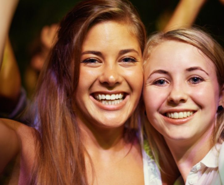 Two young girls smile and look ahead with their hands in the air. They're standing in a crowd at a concert.