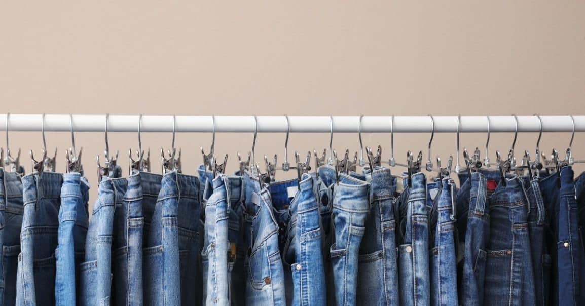 A close-up of a garment rack with various denim styles hanging on thin metal pant hangers. The garment bar is white.