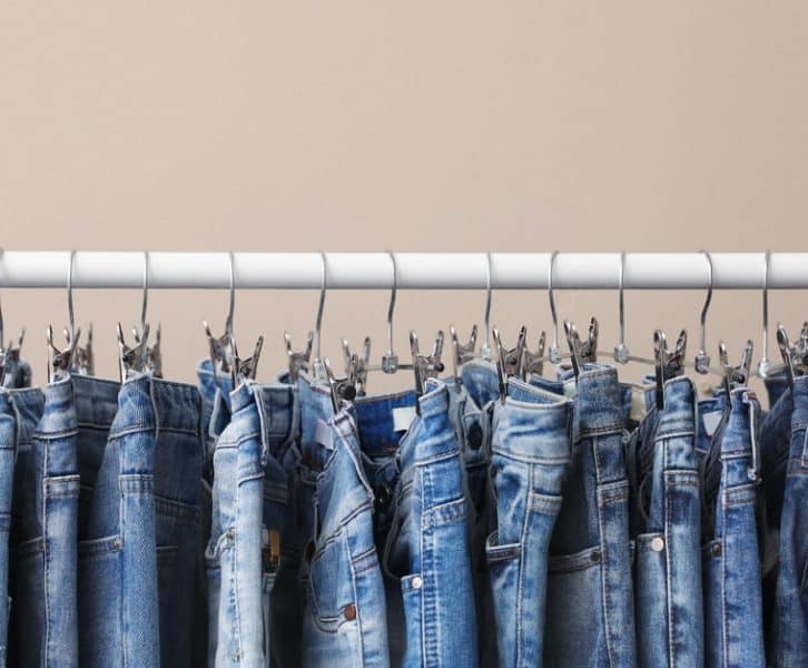 A close-up of a garment rack with various denim styles hanging on thin metal pant hangers. The garment bar is white.