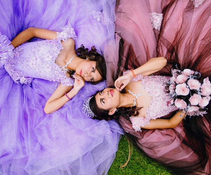 Two girls in poofy dresses, one purple and one pink, lie on the grass facing each other and making eye contact.