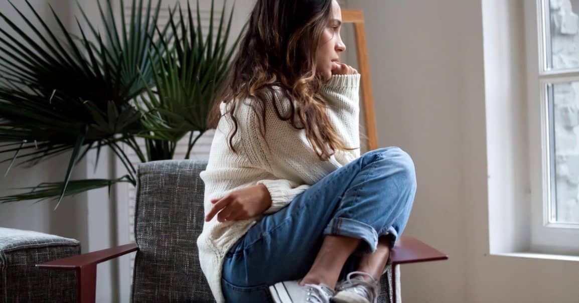A girl sitting on a lounge chair staring out the window next to her with an anxious look on her face.