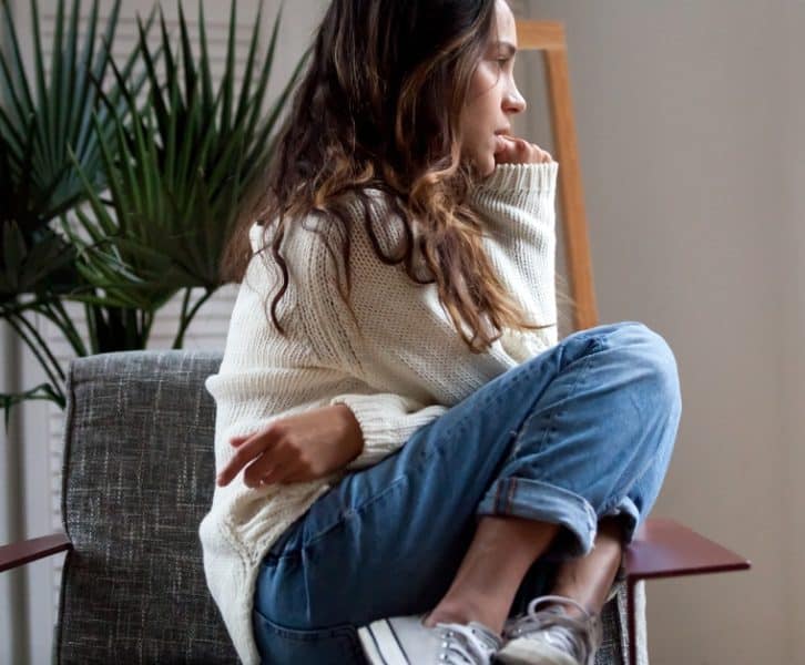 A girl sitting on a lounge chair staring out the window next to her with an anxious look on her face.