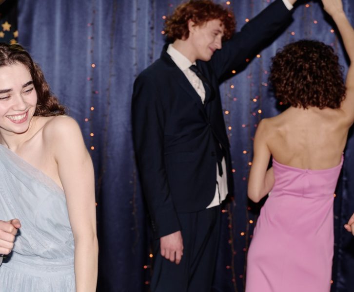 Four teenagers, including a girl in a light blue dress, dance and smile next to balloons and a blue drape backdrop with string lights.