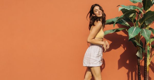 A teenage girl posing next to a tall plant against an orange wall. She wears white linen shorts and a yellow bralette-style top.