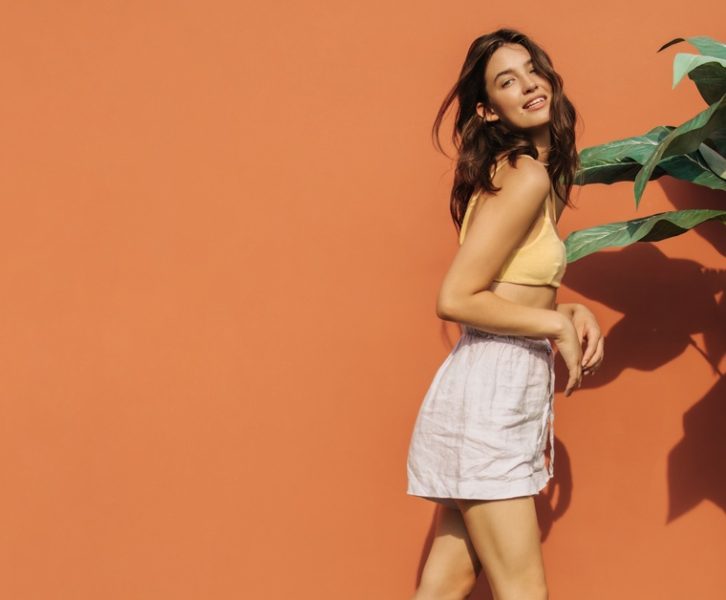 A teenage girl posing next to a tall plant against an orange wall. She wears white linen shorts and a yellow bralette-style top.