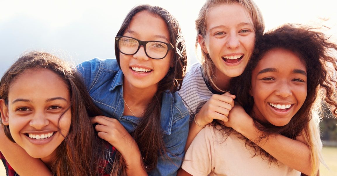 Four smiling teenage girls pause to pose for a photo. Two girls are sitting on the back of the other two girls.