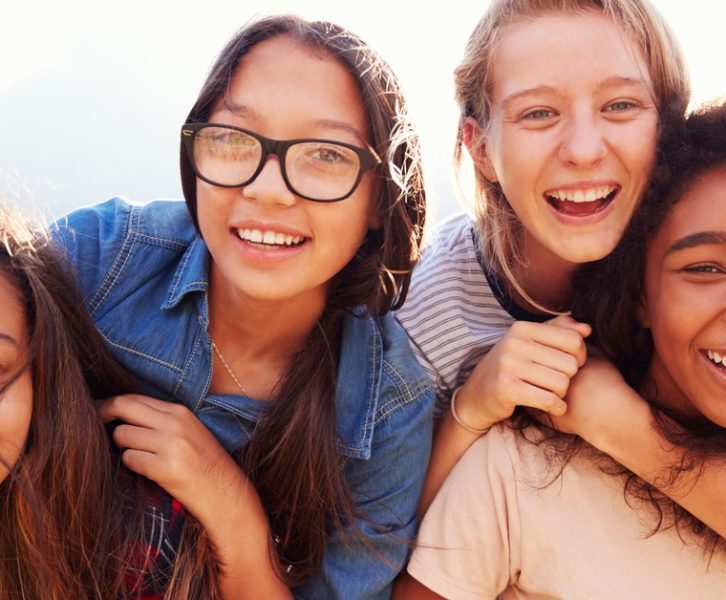 Four smiling teenage girls pause to pose for a photo. Two girls are sitting on the back of the other two girls.