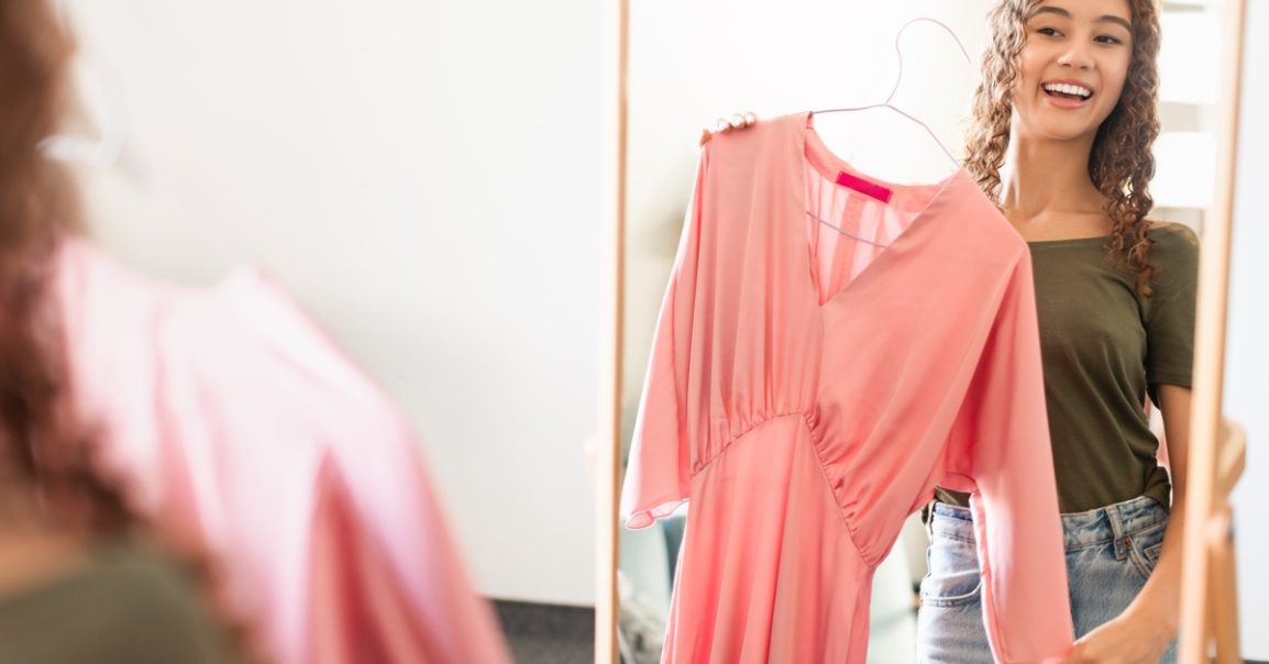 Teen girl smiles at mirror holding pink dress on hanger, trying outfit while standing in bright indoor space.