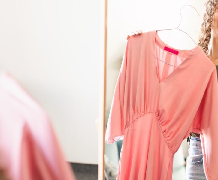 Teen girl smiles at mirror holding pink dress on hanger, trying outfit while standing in bright indoor space.