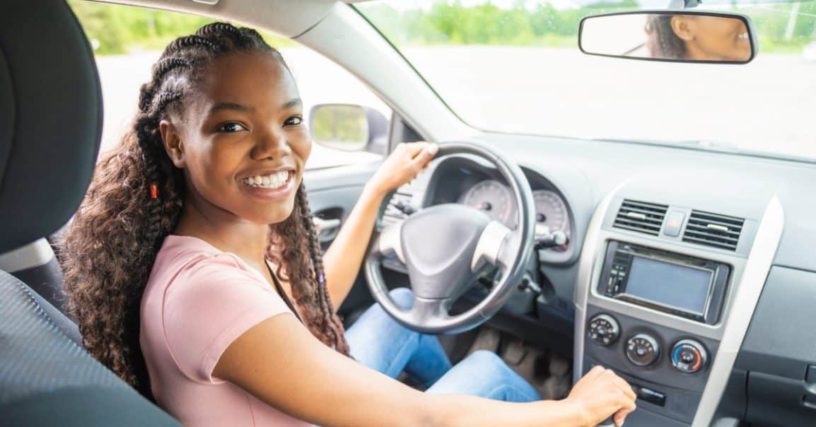 A smiling teenage girl in a pink T-shirt and jeans driving a car with her hands on the steering wheel.