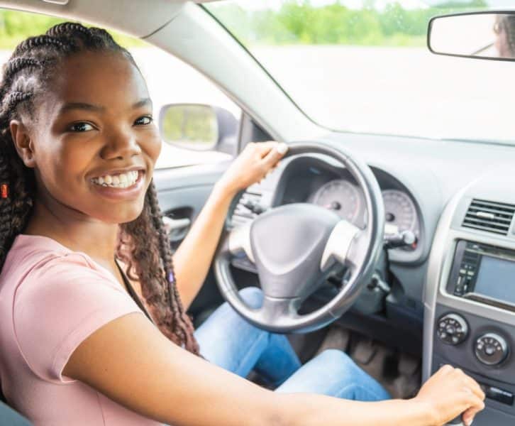 A smiling teenage girl in a pink T-shirt and jeans driving a car with her hands on the steering wheel.