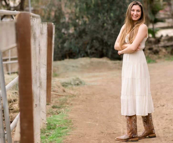 A young woman in a light dress and boots is smiling confidently while standing on a rural path near a fence.
