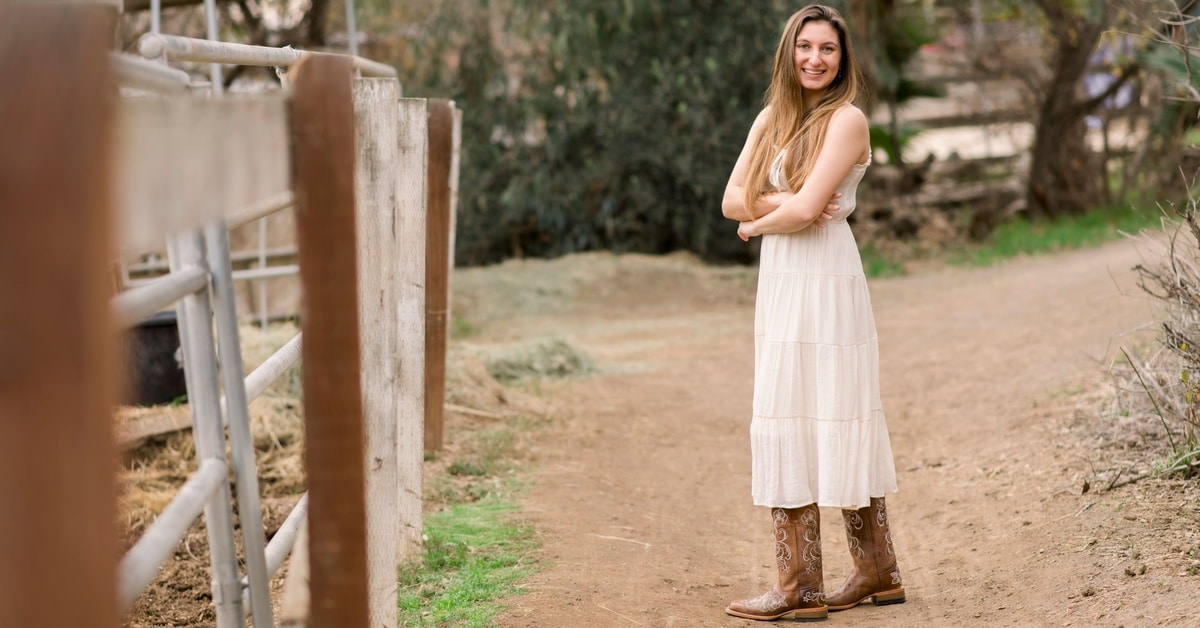 A young woman in a light dress and boots is smiling confidently while standing on a rural path near a fence.
