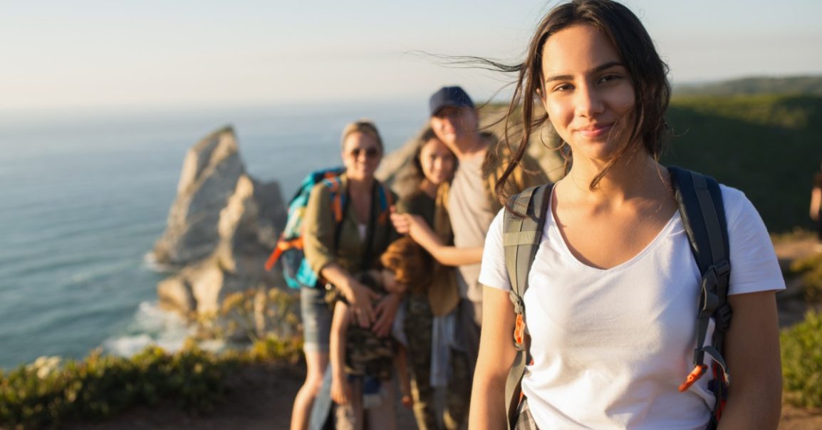 A young woman wearing a backpack standing on a coastal trail, with four blurred people behind her near cliffs.