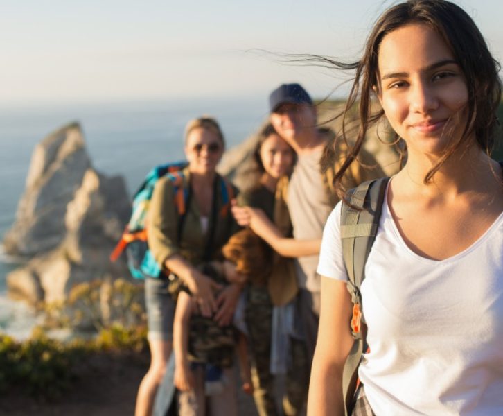 A young woman wearing a backpack standing on a coastal trail, with four blurred people behind her near cliffs.
