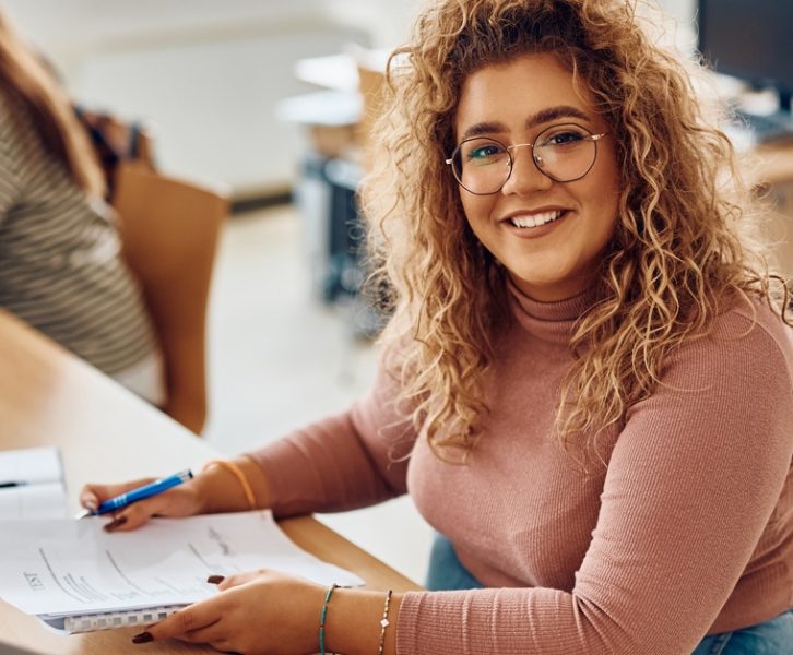 A smiling young woman sits at a wooden table holding a notebook and pen. An open laptop sits in front of her.