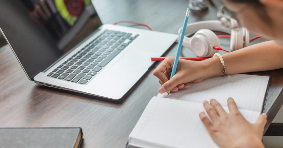 A girl writes notes in a notebook beside a laptop and headphones while studying or working at a desk.