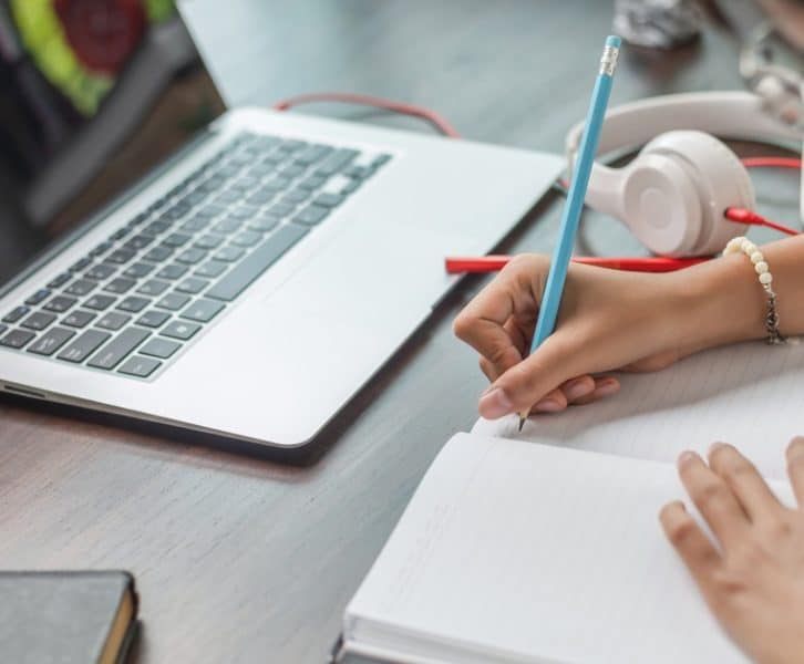 A girl writes notes in a notebook beside a laptop and headphones while studying or working at a desk.