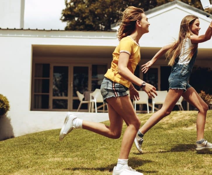 Two girls run across a grassy yard while one holds a toy airplane, with a house and patio in the background.