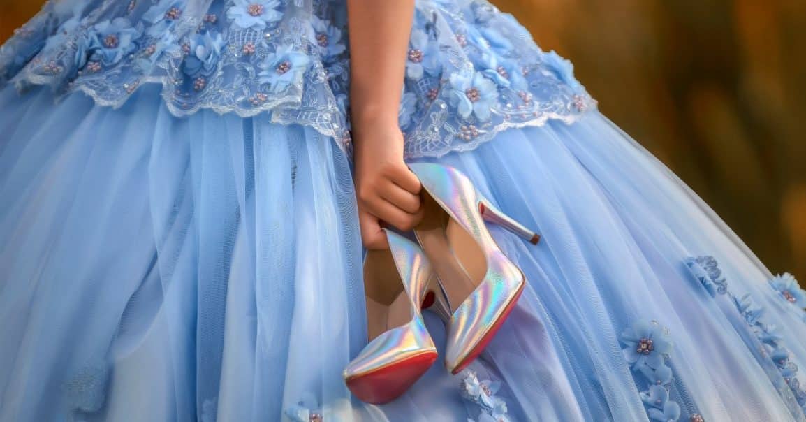  Hand holding iridescent high heels beneath a blue embroidered ball gown with layered tulle and floral appliqués.