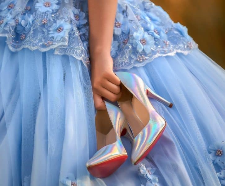  Hand holding iridescent high heels beneath a blue embroidered ball gown with layered tulle and floral appliqués.