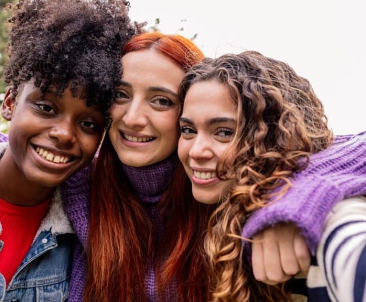 Three teenage girls smile together outdoors as one holds up a phone to take a selfie with her friends.