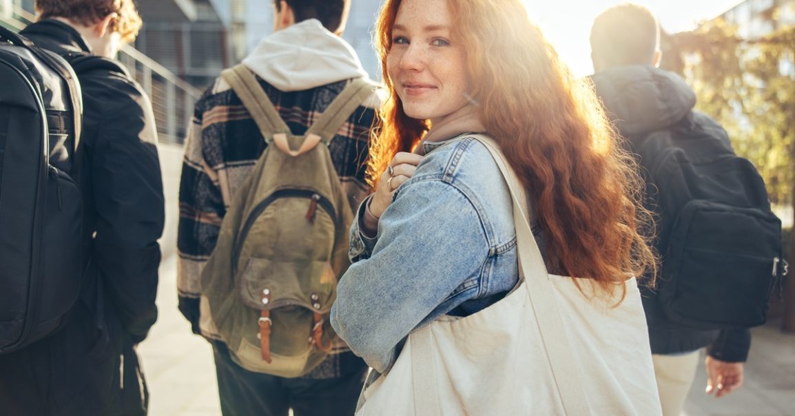 A teenager smirks with the sun behind her and high schoolers in front of her. She has long red hair.