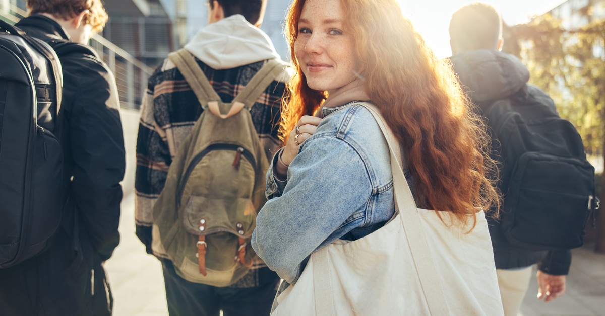 A teenager smirks with the sun behind her and high schoolers in front of her. She has long red hair.