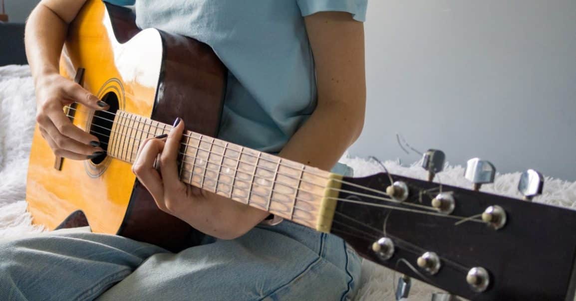 A close-up of a teenager wearing a blue shirt and jeans sitting on a bed while playing an acoustic guitar.